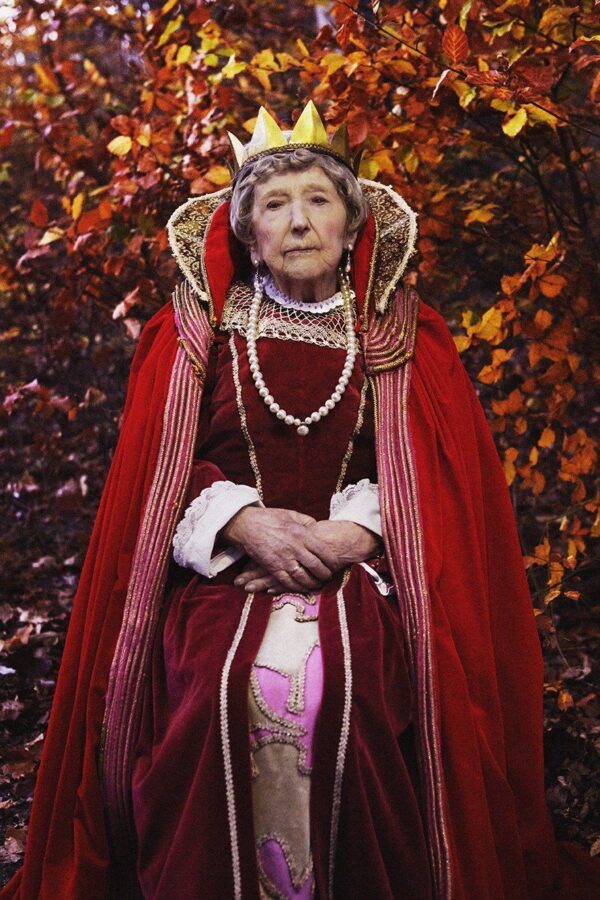 Dagny Carlsson 107 år med guldkrona på huvudet, drottning likt sitter hon i mörkröd klänning, pärlhalsband med en klarröda mantel över axlarna med händerna knäppta i knät. Omgiven av höstlövens färger